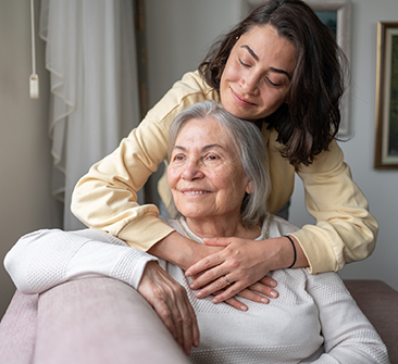 A smiling young woman stands behind her elderly mother giving her a hug