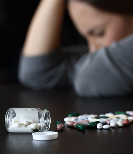 A woman rests her head on a counter next to a pile of pills