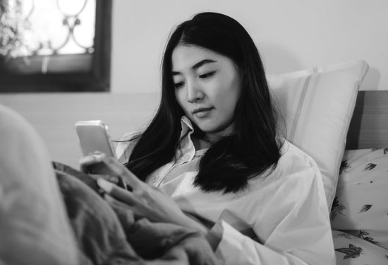 A woman sits on her bed and looks at her phone.