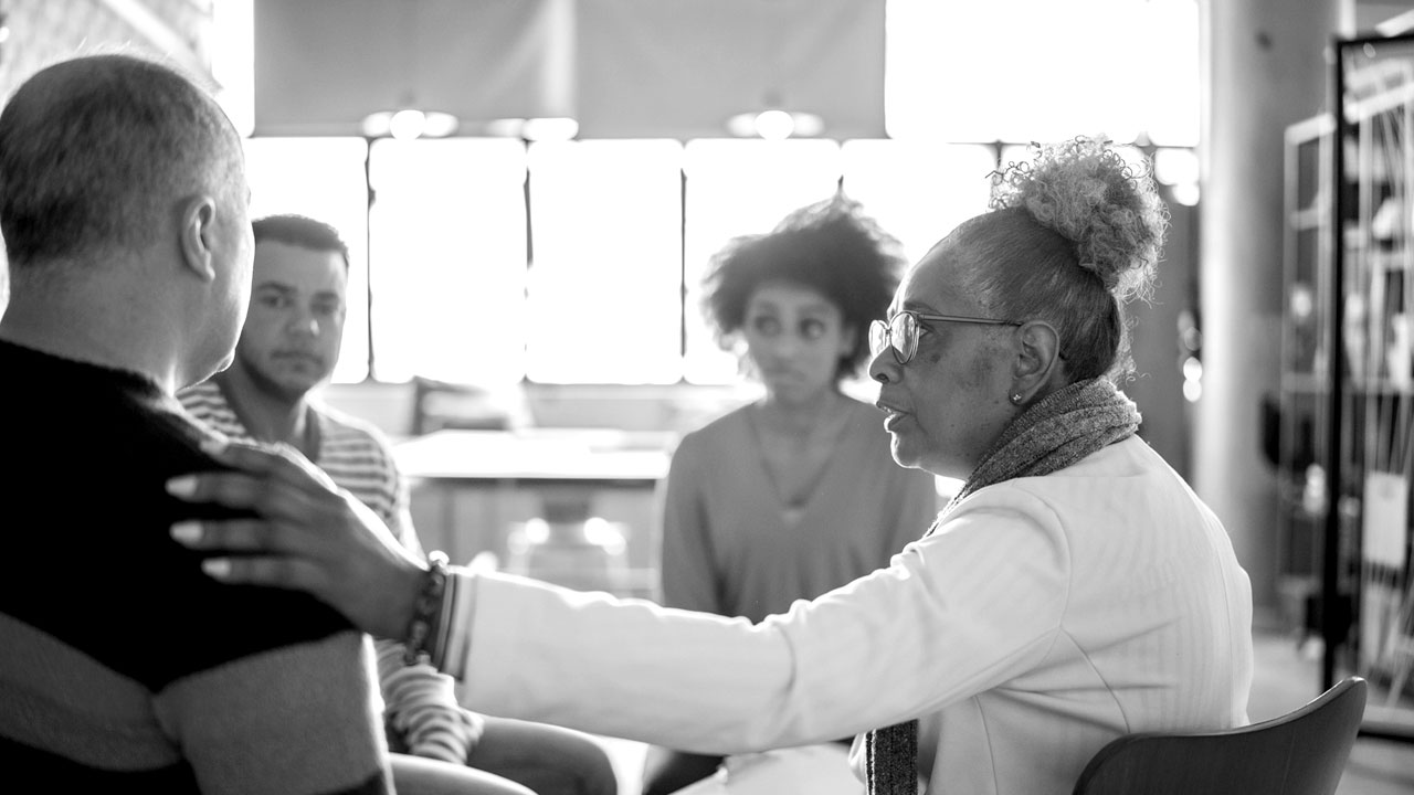 A woman puts her hand on a person's shoulder as two other people watch in the background.