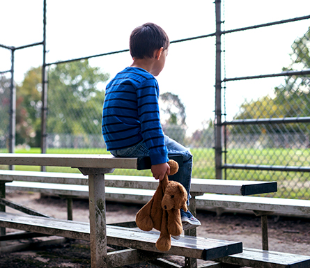 A young boy sitting in bleachers with his back to the camera, holding a stuffed animal