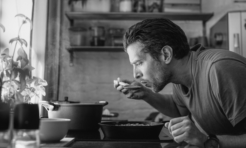 A man sips stews as he cooks at a stovetop.