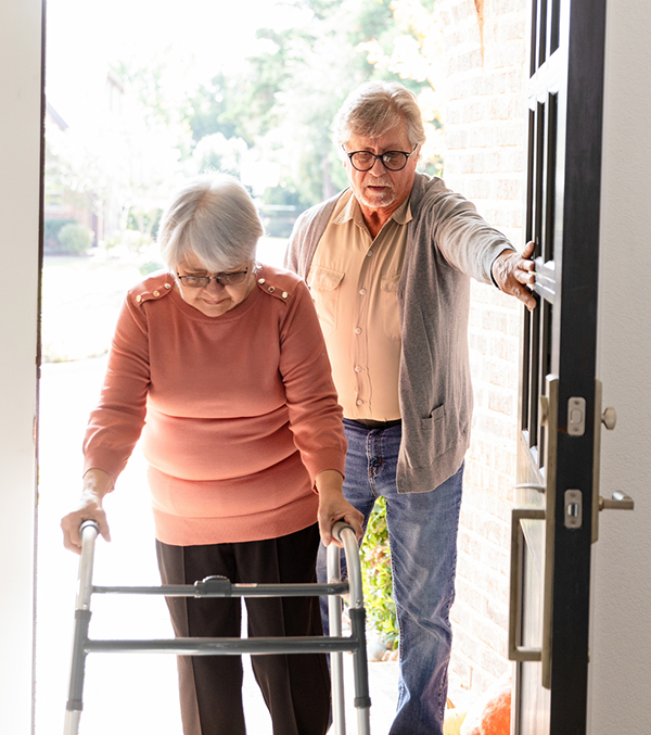 A woman uses a walker to enter her home while a man holds the door open.