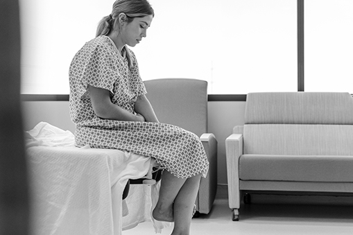 A woman wearing a medical gown sits on the edge of a table.