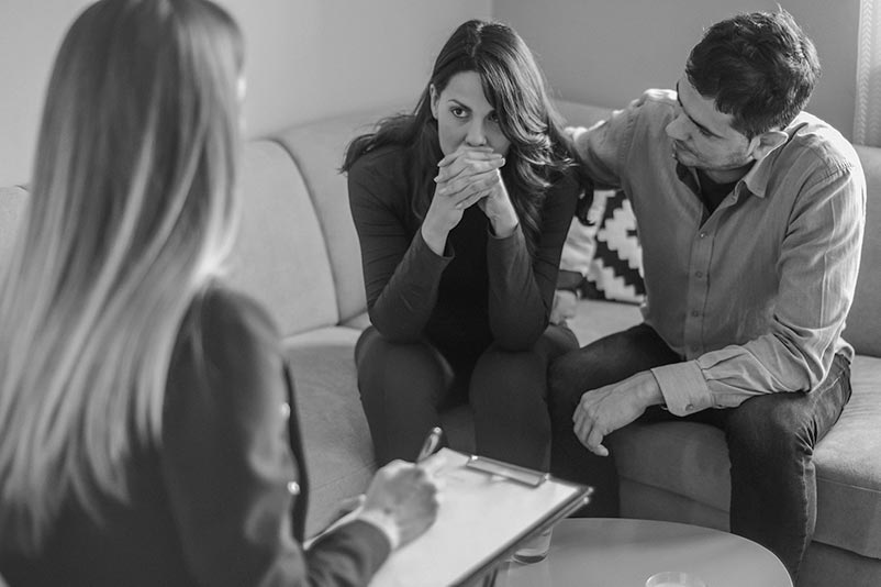 A woman folds her hands in front of her face as a man to her right puts his arm on her shoulder and another woman talks to her.