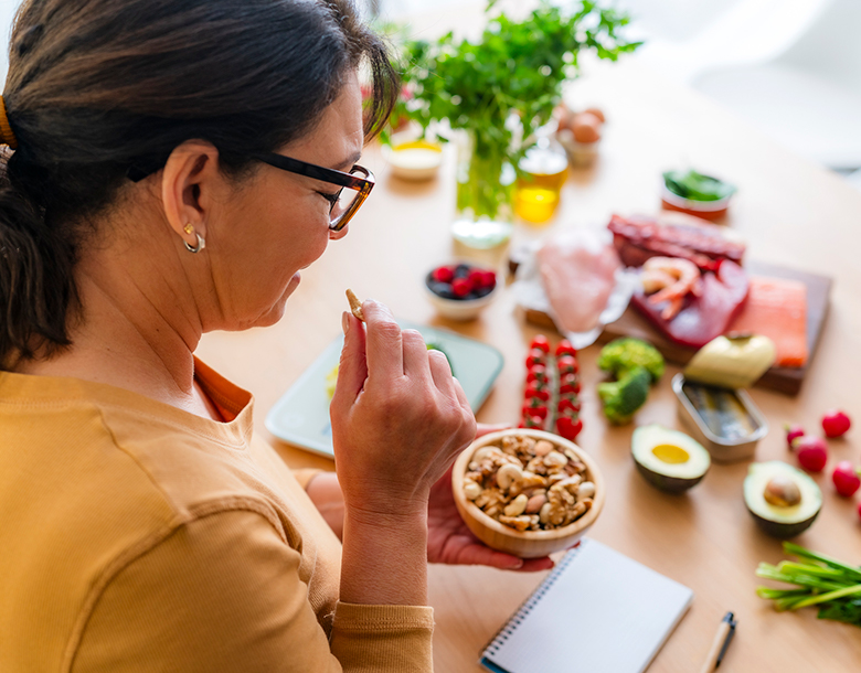 A woman eats a cashew with a notebook and various healthy foods laid out before her.