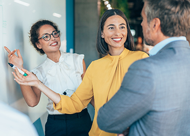 Two women point to a whiteboard during a meeting in a conference room while a man stands with his arms crossed. 