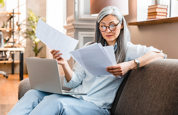 A woman looks over some paperwork while sitting with her laptop.