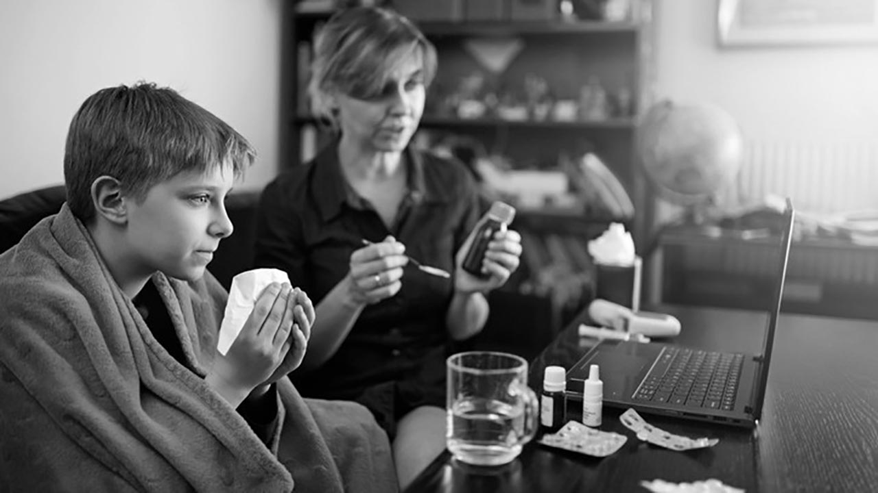 A boy blows his nose while his mother gets medicine ready