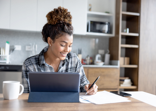 A woman takes notes on some paper next to her tablet.