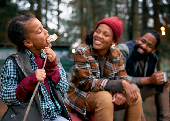 A young girl takes a bite out of a smore at a campsite while her parents look on.