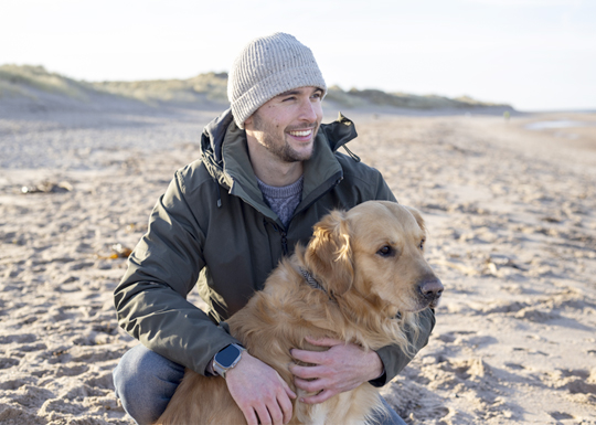 A young man holds a golden retriever on the beach while looking off into the distance.