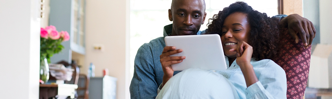 A pregnant woman smiles while talking on her tablet. Her partner sites next to her.