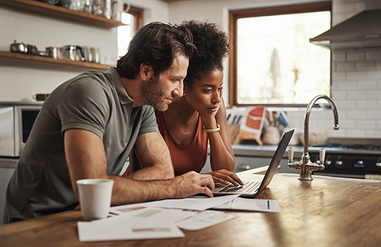 A man and a woman look over their benefits on a laptop while standing at the kitchen counter.