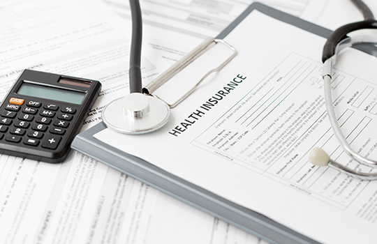 Health insurance forms on a clipboard lie next a stethoscope and a pocket calculator.