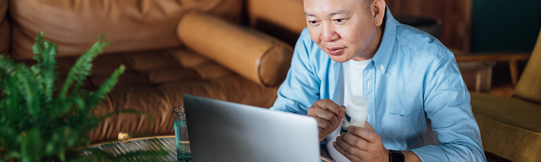 A man sits reading what's on his laptop while holding a bottle of prescription medication.
