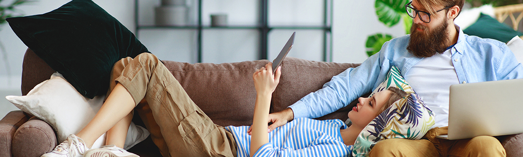 A woman lies on a couch looking at her tablet with a man sitting at her side.