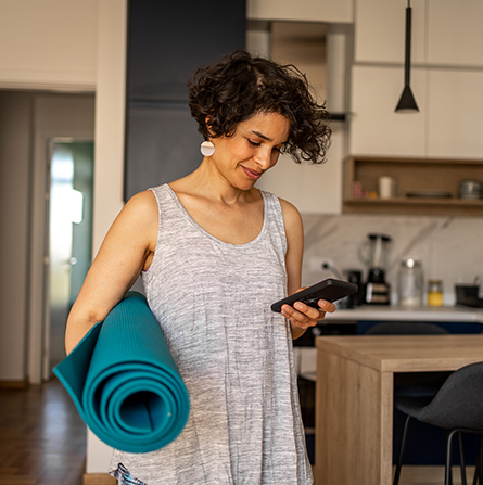 A woman stands holding a yoga mat while scrolling on her phone.