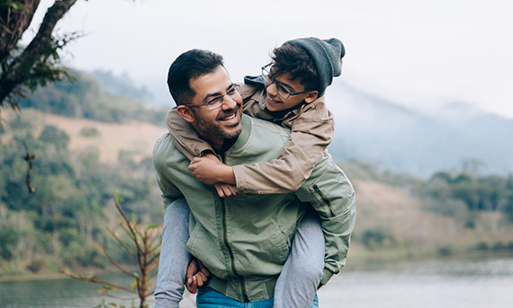A man carries his son on his back as they smile and walk down a path together.