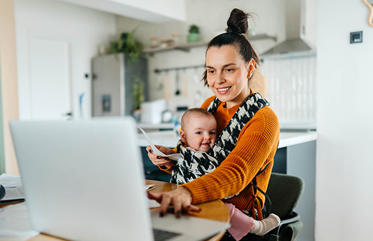 A woman holds a baby and works on her laptop.