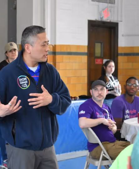 A man speaks to a group of people gathered in a gymnasium.