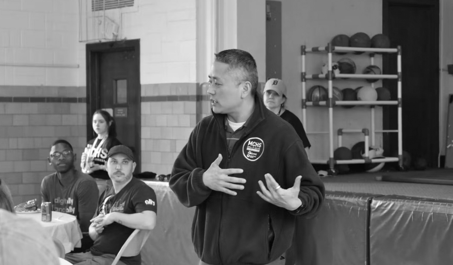 A man speaks to a group of people gathered in a gymnasium.