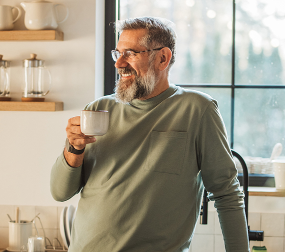 An older man stands in his kitchen with a cup of coffee.