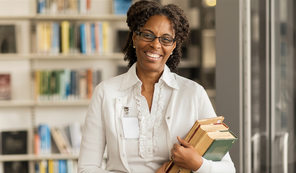 A smiling woman stands while holding some books in a library. 