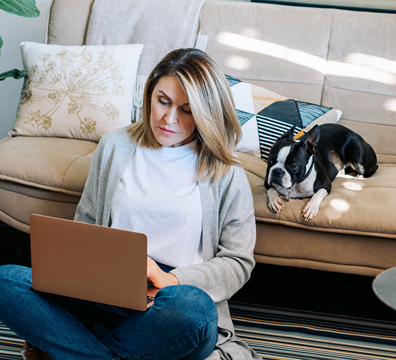Woman sitting on the floor looking at her laptop with a dog lying on the couch behind her