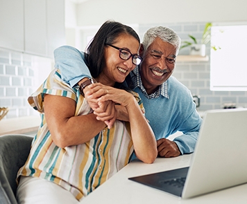An older couple sits smiling at a computer with their arms around one another