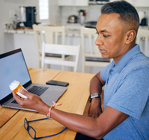 A man looks at a prescription bottle while sitting in front of his laptop.