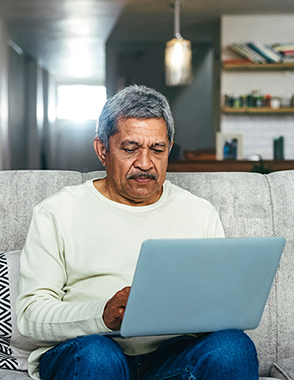 A senior man types on his laptop while sitting on a couch.