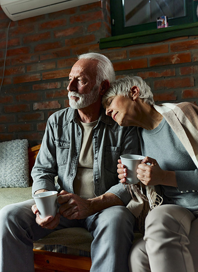 An older woman rests her head lovingly on an older man as they drink coffee.