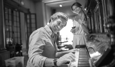 A man plays piano as a woman looks on in the background.