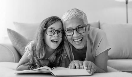 A woman and a child, each with glasses, smile as they look up from a book they're reading together.