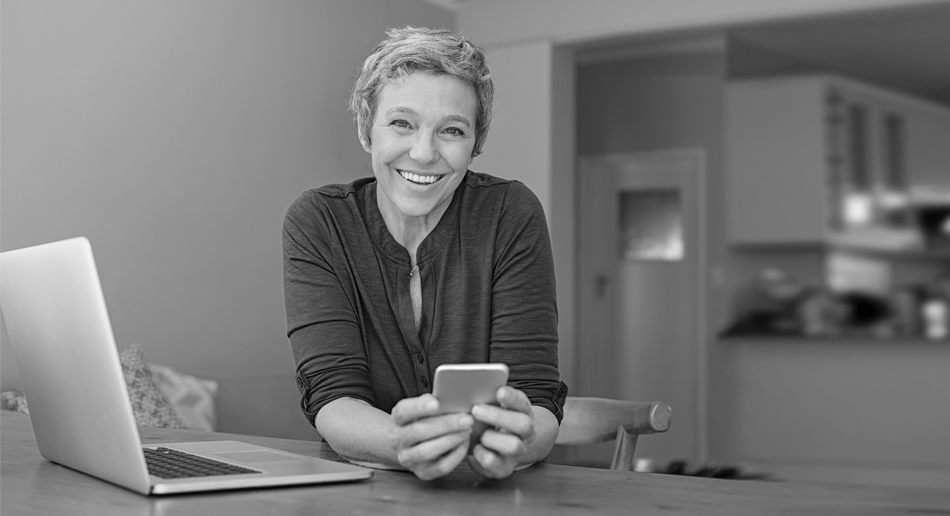 A woman smiles while holding a smartphone with an open laptop next to her.