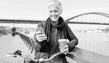 A woman with a bike stops to check her smart phone while holding a coffee.