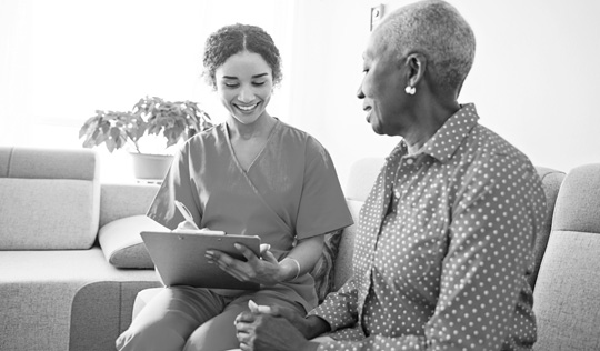 A woman in scrubs looks at a clipboard while she speaks to a patient.