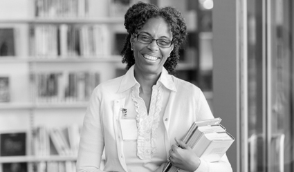 A smiling woman stands while holding some books in a library. 