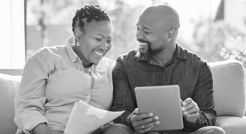 Happy couple sitting on couch, looking at tablet and paper together.