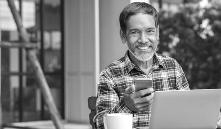 A man sits at his computer and smiles while checking his smartphone.