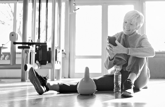 A woman smiles, sits on the floor and looks at her phone after working out.