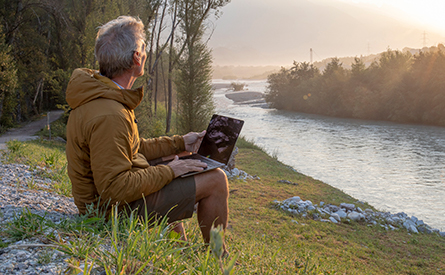 A man sits on a riverbank with his laptop open.