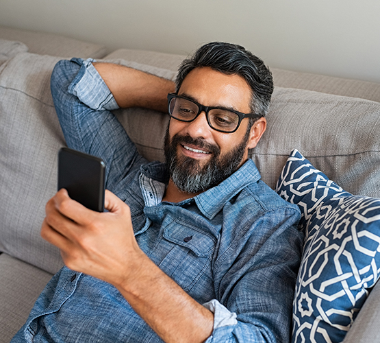 A man leans back on a couch while looking at the BCBSM mobile app on his smartphone.
