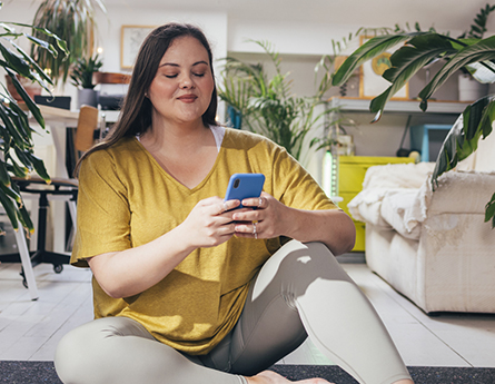 A woman surrounded by houseplants sits on the floor and looks at her smartphone.