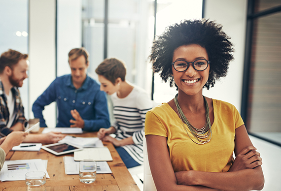 A smiling woman stands in front of a group of people collaborating on a project.
