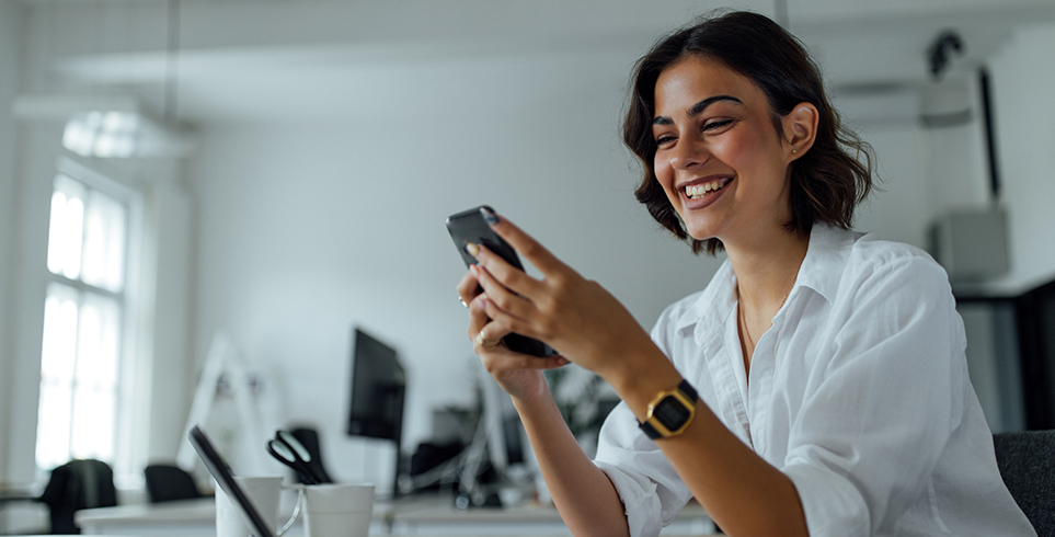 A woman seated at a desk smiles while looking at her smartphone.