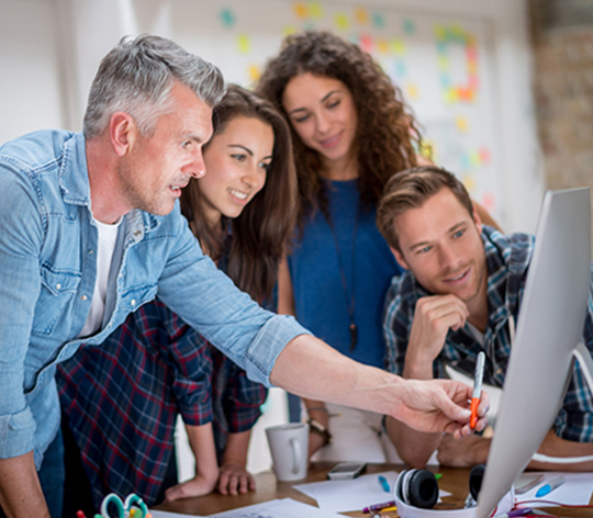 A group of coworkers stand in front of a computer monitor and discuss a project together.