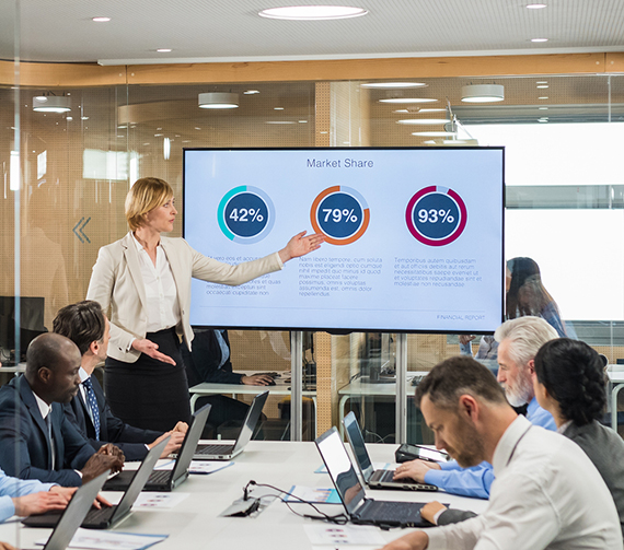 A woman gestures at a PowerPoint presentation in a conference room full of people.