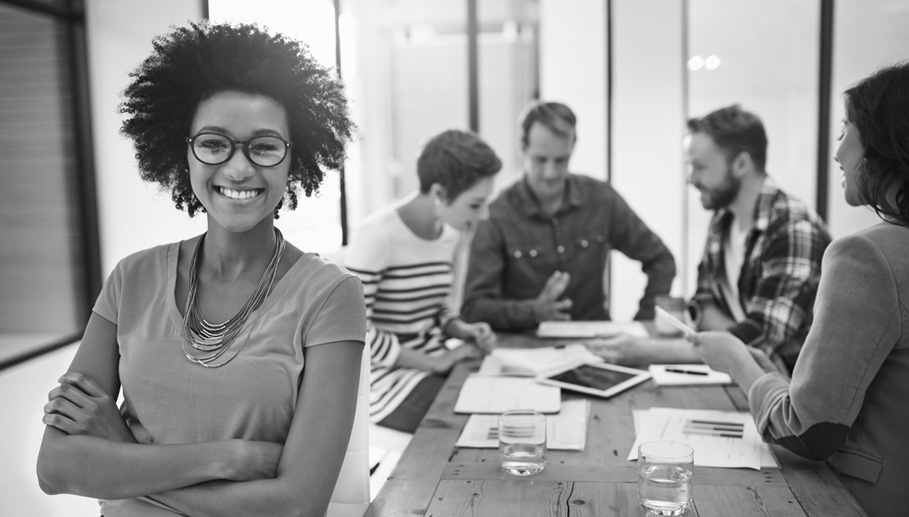 A smiling African-American woman stands in front of a group of people collaborating on a project.